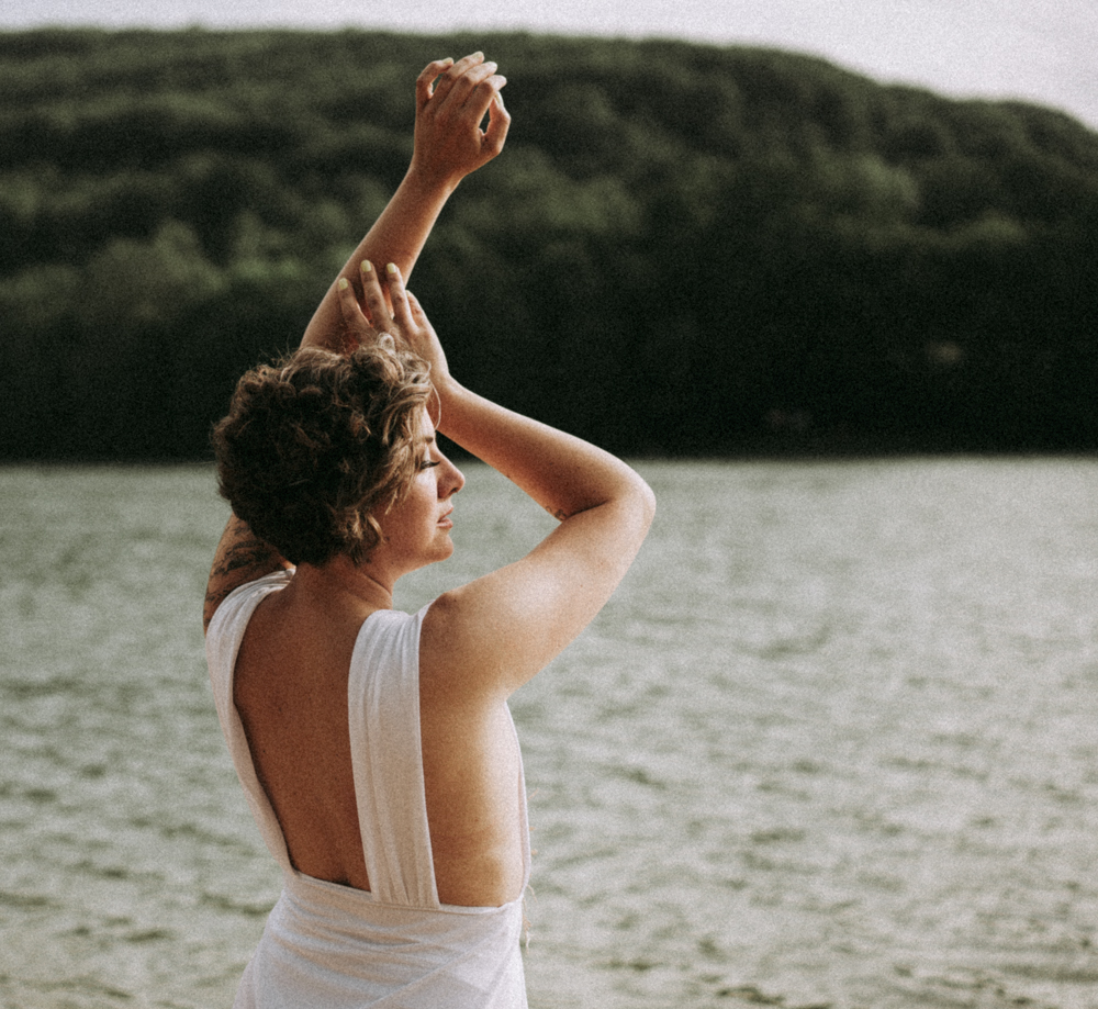 Femme posant gracieusement au bord d’un lac, vêtue d’une robe blanche au dos ouvert, en pleine nature apaisante.